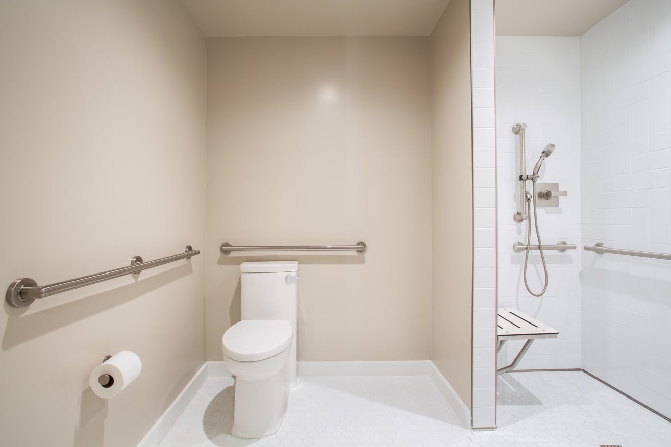 A toilet and a shower stall surrounded by hand rails in a large open and accessible bathroom area.