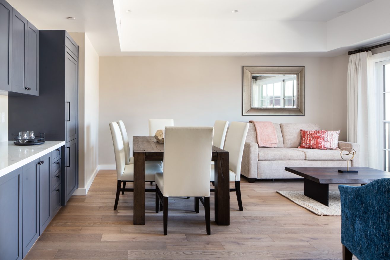 A guest room with beige chairs at a dark wood dining table. And a beige love seat with red pillows behind a dark wood coffee table.