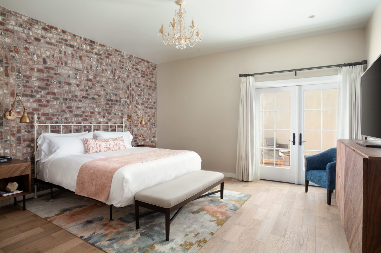 A bed with a bench at the foot of it and chandelier lighting above it. A brick wall with bedside lighting is visible behind the headboard.