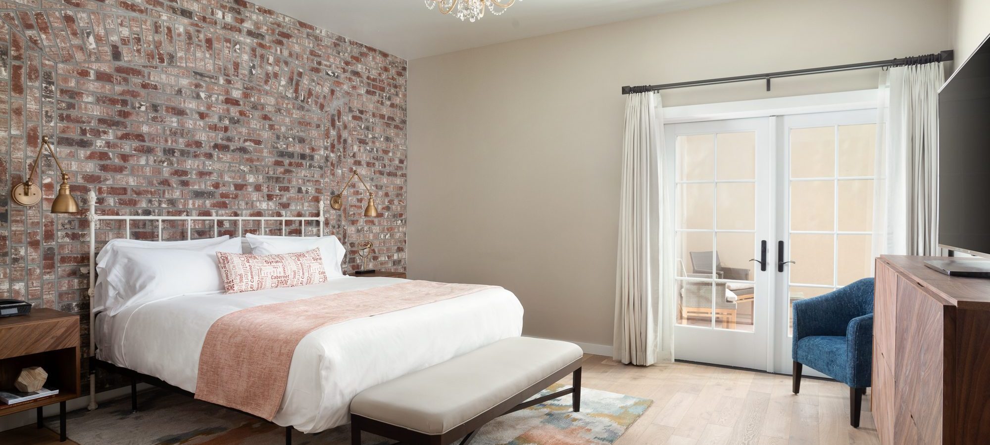 A bed with a bench at the foot of it and chandelier lighting above it. A brick wall with bedside lighting is visible behind the headboard.