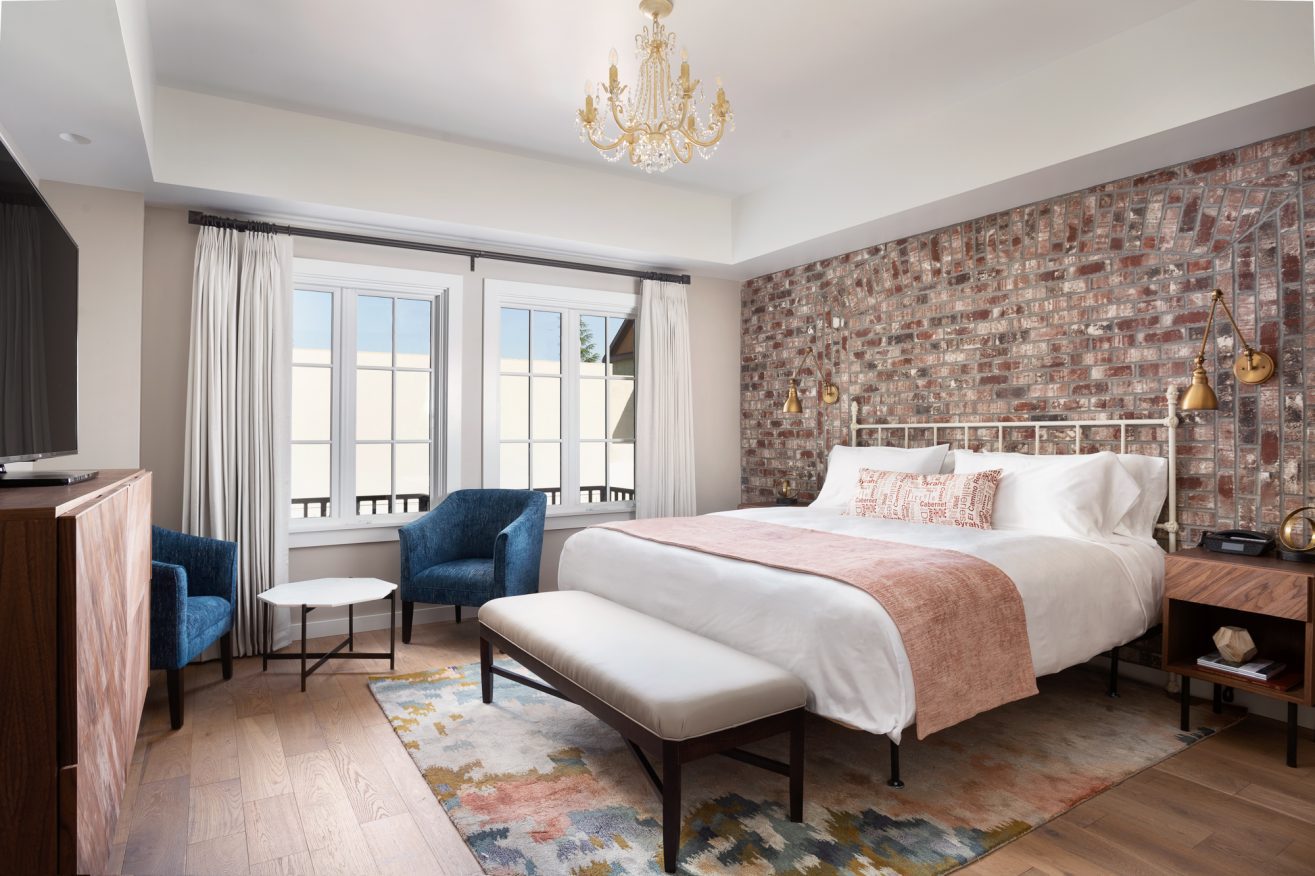 A bed with a bench at the foot of it and chandelier lighting above it. A brick wall with bedside lighting is visible behind the headboard.