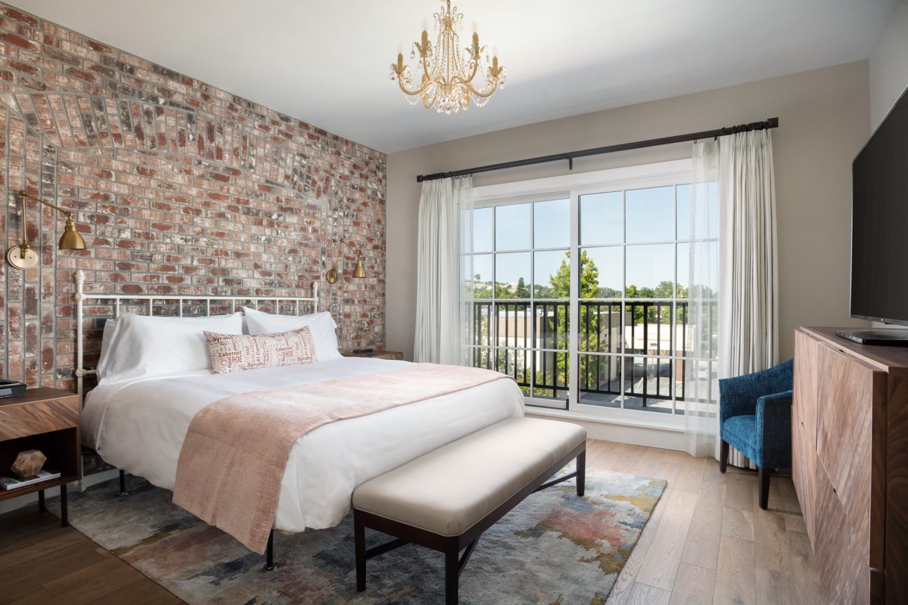 A king bed with white blanket and pink accents with a bench at the foot of the bed, Large glass doors overlook greenery