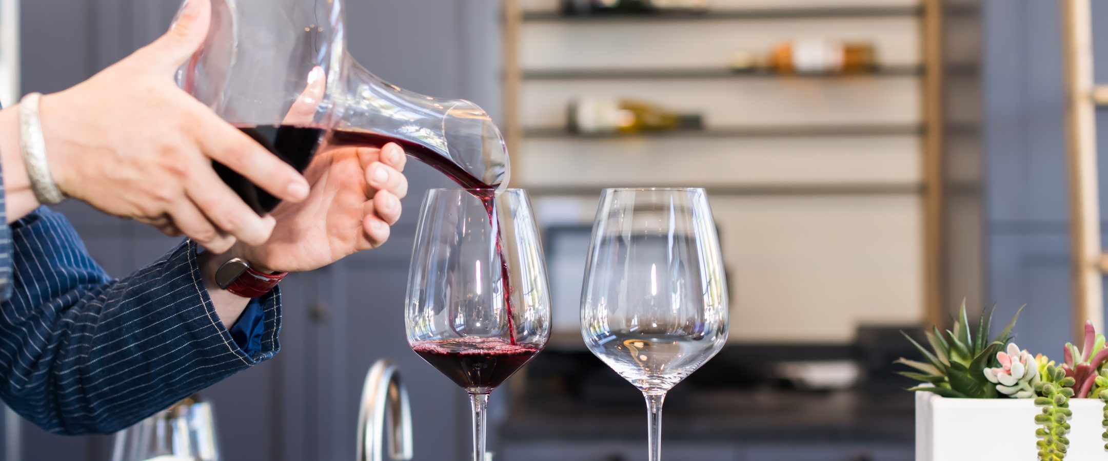 Close up of hands holding a decanter pouring red wine into wine g lasses on a bar. A rack with wine is in the background.