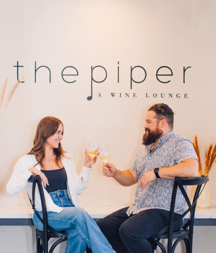 A woman and a man sit on bar stools enjoying wine. The Piper wine lounge sign is on the wall behind them.