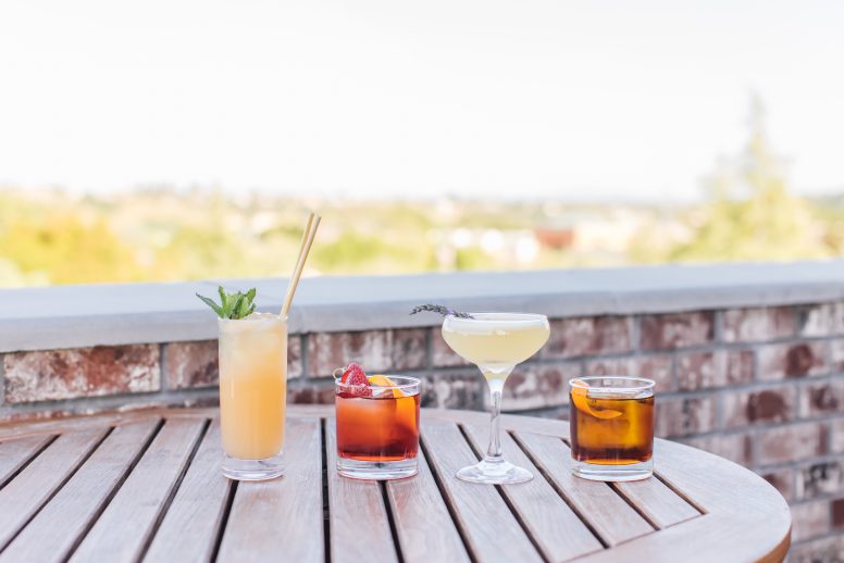 A variety of cocktails in different glasses on a wooden table on a rooftop. The view of greenery is behind the glasses.