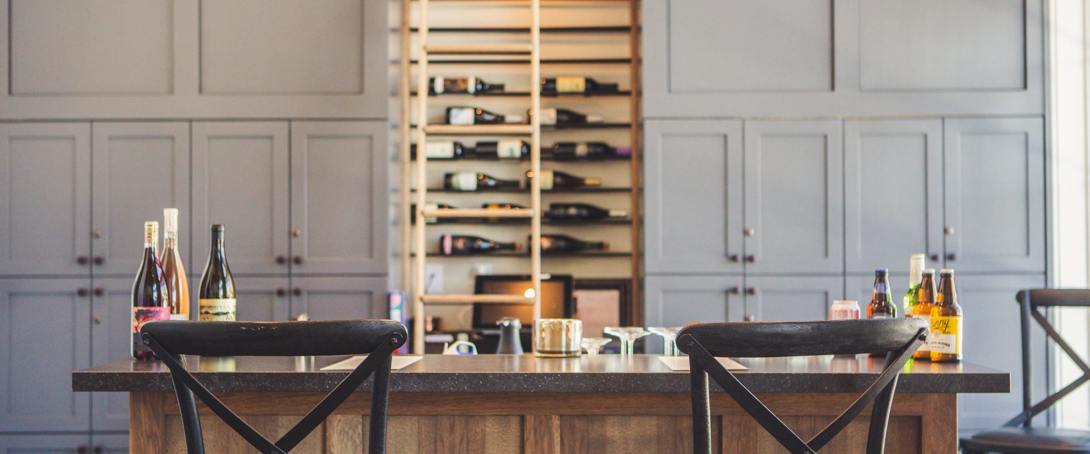 A bar with two chairs in front, bottles of wine and other beverages on either side. A wine rack with bottles and a ladder is behind the bar.