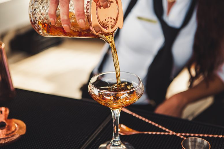 A close up of a bartender pouring a cocktail from a crystal cylinder into a coupe glass.