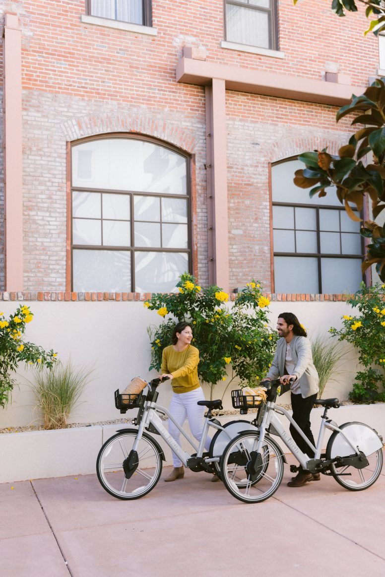 A woman is a green sweater standing beside a bicycle holding the handlebars. She is standing in front of a brick building with big arched windows.