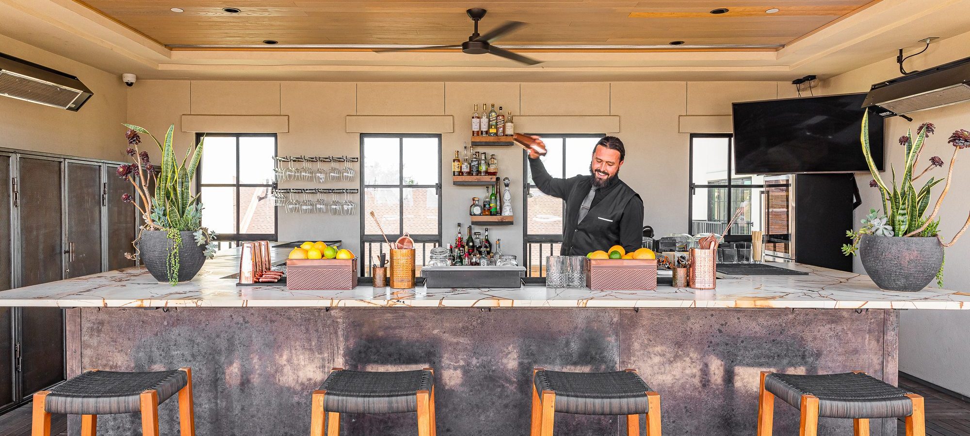 A bartender stands behind a bar shaking a cocktail. THree barstools are in the foreground