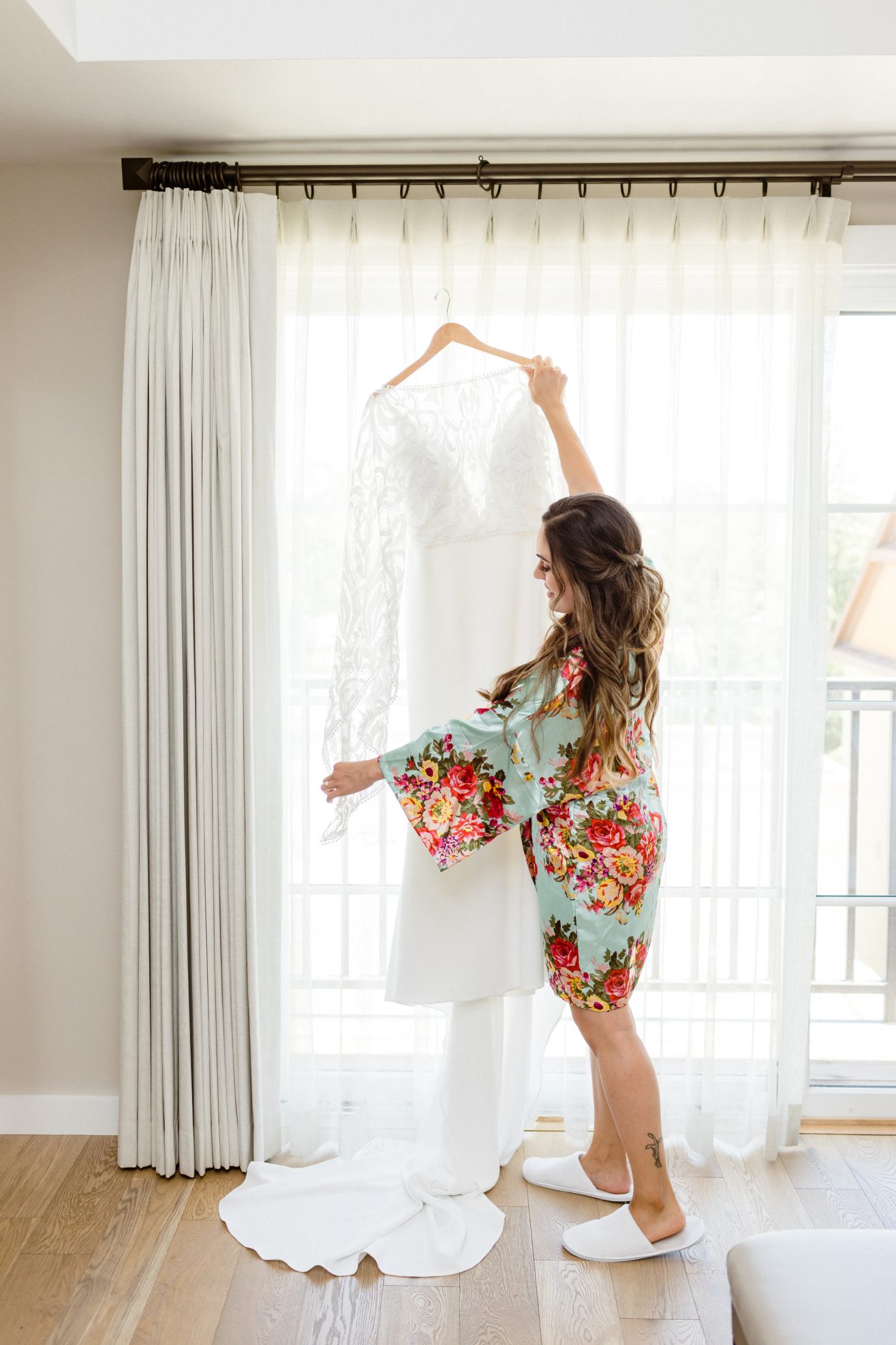 A girl in a colorful robe holding up a white wedding dress in front of white curtains in guest room.