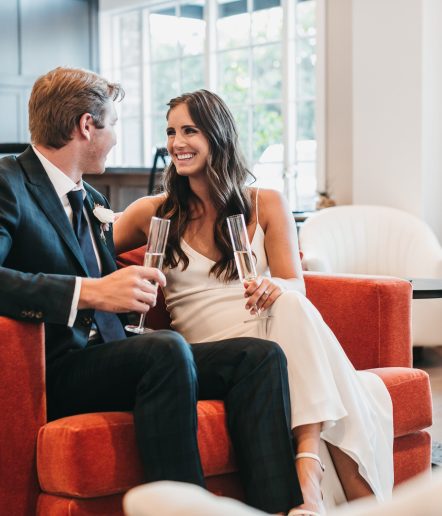 A bride and groom seated on a red sofa facing each other and holding champagne glasses.