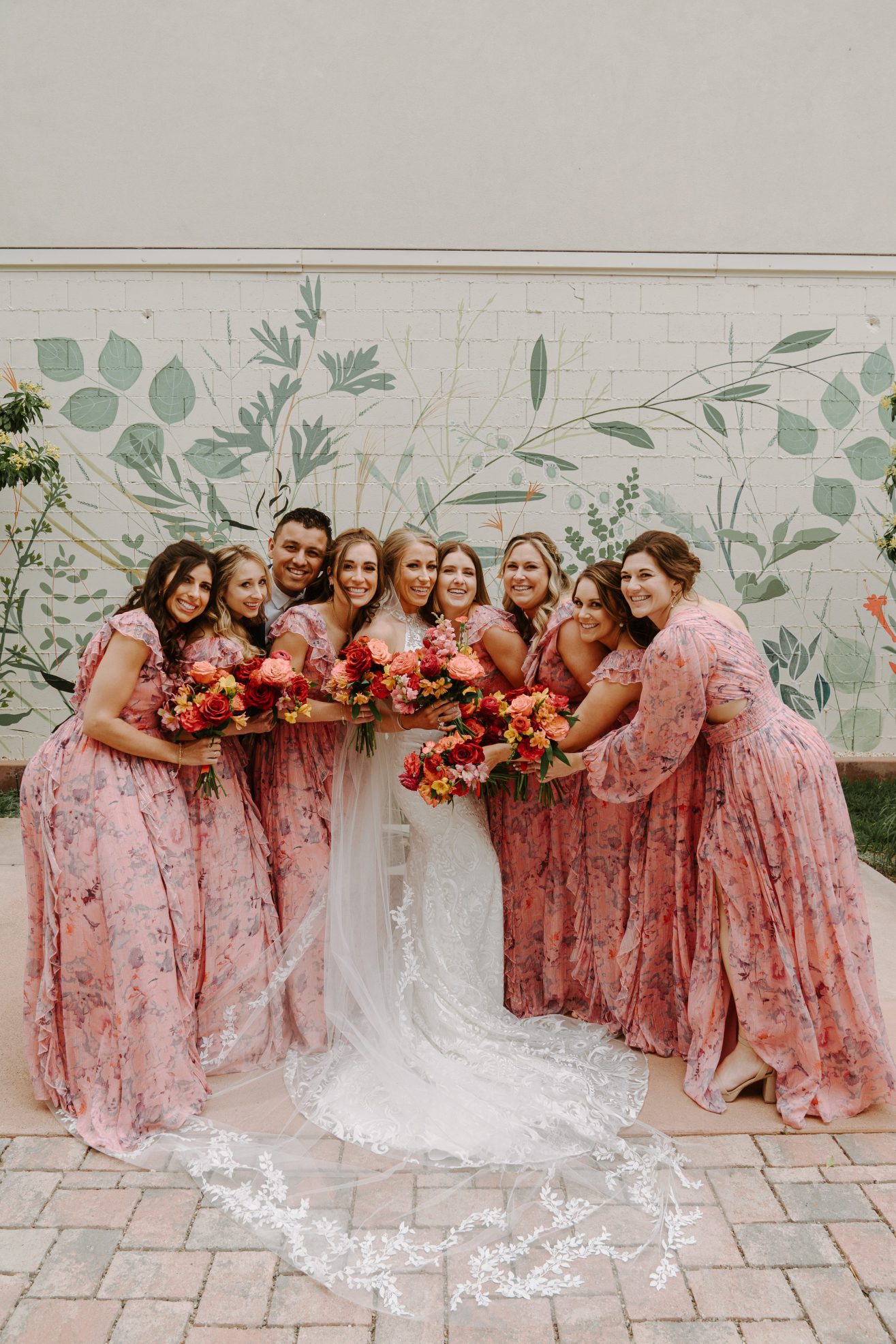 A bride and groom cheerfully posing together with the bridesmaids while holding flower bouquets.