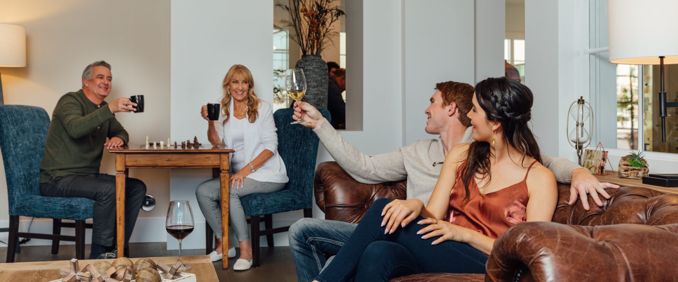 Two couples in a lounge. The older couple hold coffee cups while sitting at a chess table. The younger couple sits on a sofa. the man is holding a glass of white wine.