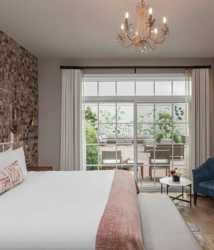 A king bed with white blanket and pink accents with a bench at the foot of the bed, Large glass doors overlook greenery