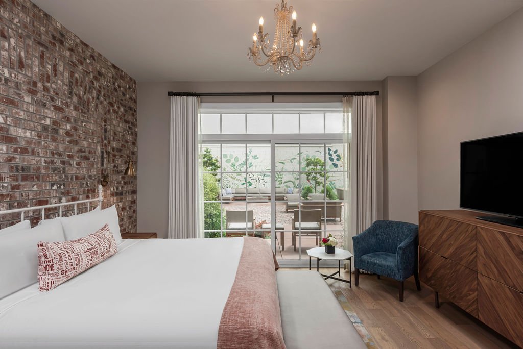 A king bed with white blanket and pink accents with a bench at the foot of the bed, Large glass doors overlook greenery