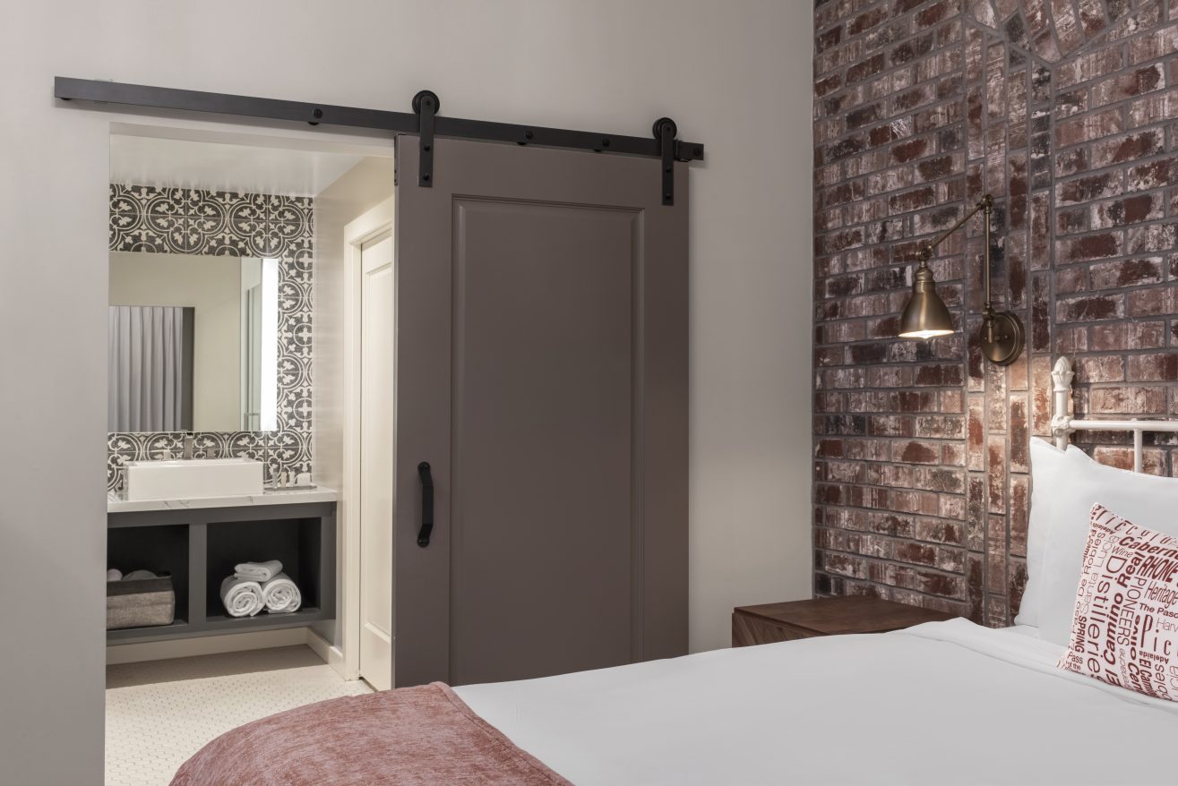 Part of a bed is showing in a room with a barn door to a bathroom.