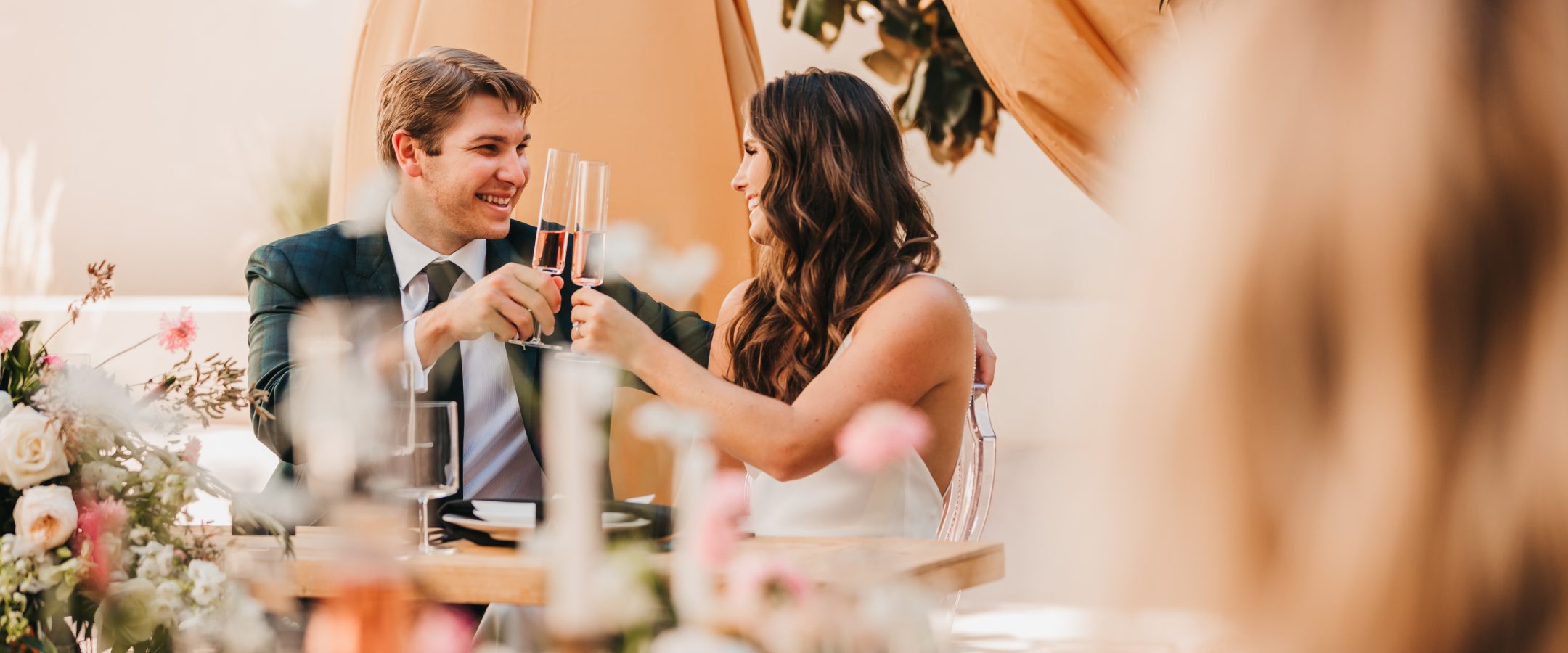 A bride and groom seated facing each other at a dinner banquet, holding their champagne glasses up together.