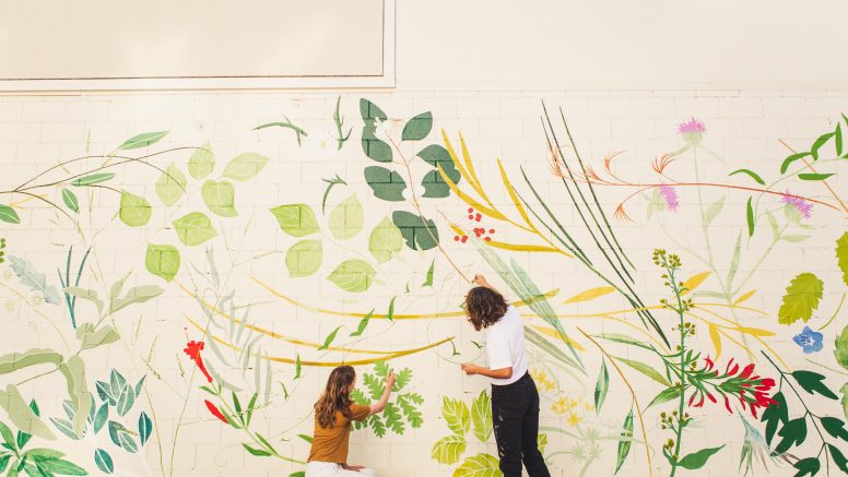 Two woman painting a large floral mural on a side of a white building