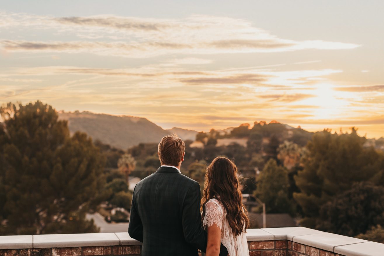 A bride and groom looking towards the sunset view from a rooftop outdoors.