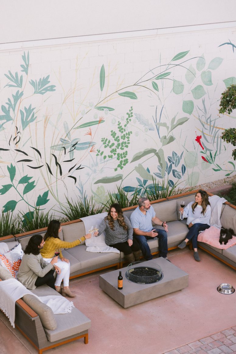 a group of people sitting at a sectional patio set around a firepit with a flora mural behind them