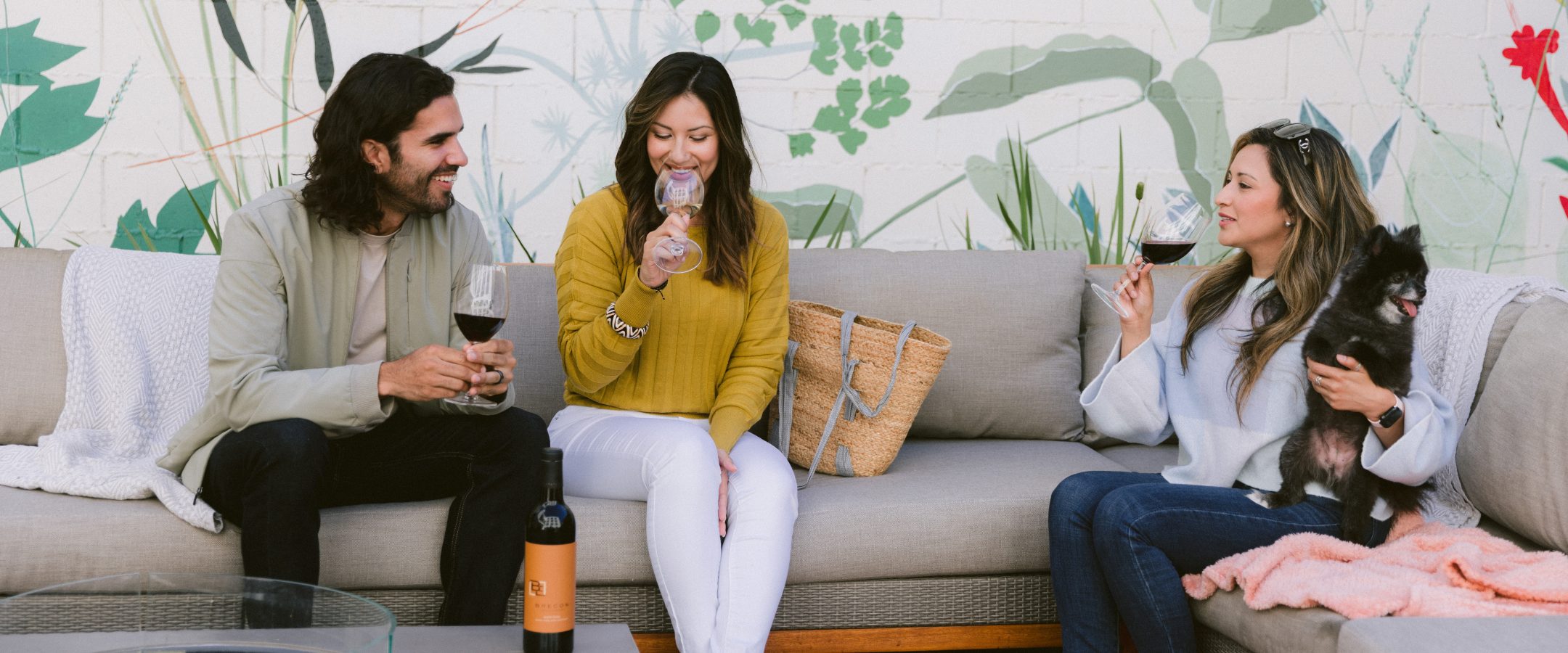 A man, two ladies and a small dog sitting out while enjoying glasses of wine together.